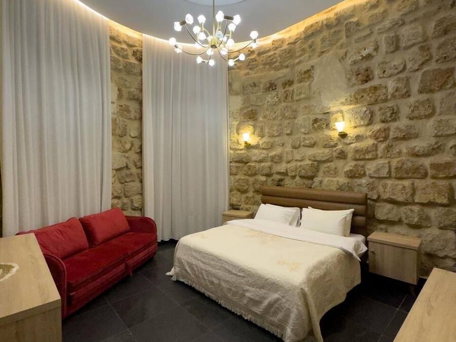 Un dormitorio con pared de piedra, una lámpara redonda en el techo, una cama con ropa blanca, un sofá rojo y una mesita de madera.