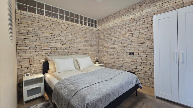 A bedroom with a stone wall, a bed with a grey quilt, a white wardrobe, and a skylight.