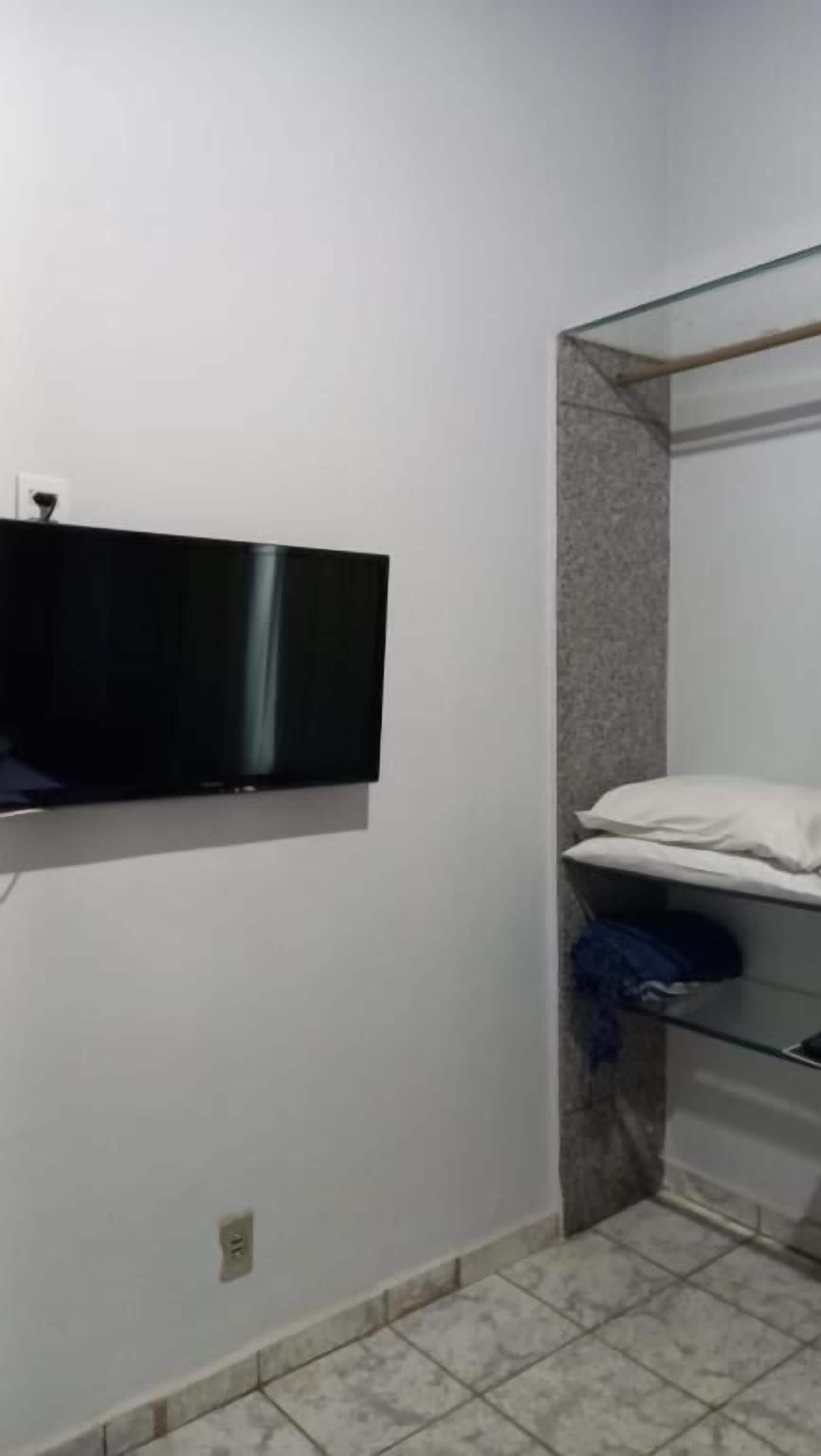 Quarto pequeno, com poucos móveis, onde se vê uma televisão na parede, chão de azulejos e um canto com prateleira de vidro, com um travesseiro branco e uma peça de roupa azul dobrada.