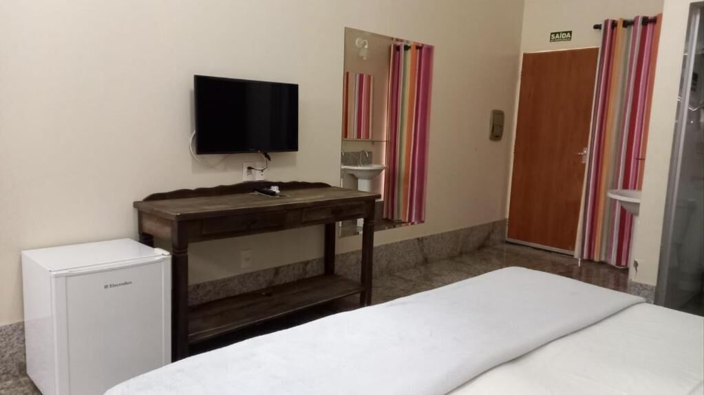 A hotel room with a bed, a small desk with a television, a refrigerator, a bathroom with a mirror, and a door marked "SAÍDA".