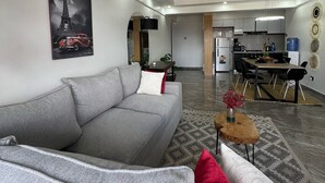 Een moderne woonkamer met een grijze bank, een rode kussen en een salontafel. Achtergrond is een keuken met een eettafel en stoelen.