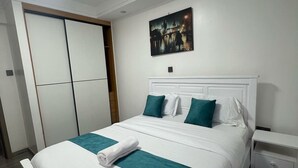Een net opgemaakt bed met wit en teal beddengoed, een schilderij van een stadsgezicht aan de muur, en een witte kledingkast met schuifdeuren.
