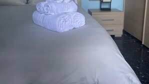Un letto rifatto con asciugamani bianchi arrotolati, una testiera in legno, un comodino con una foto incorniciata e un quadro raffigurante una barca a vela appeso al muro.