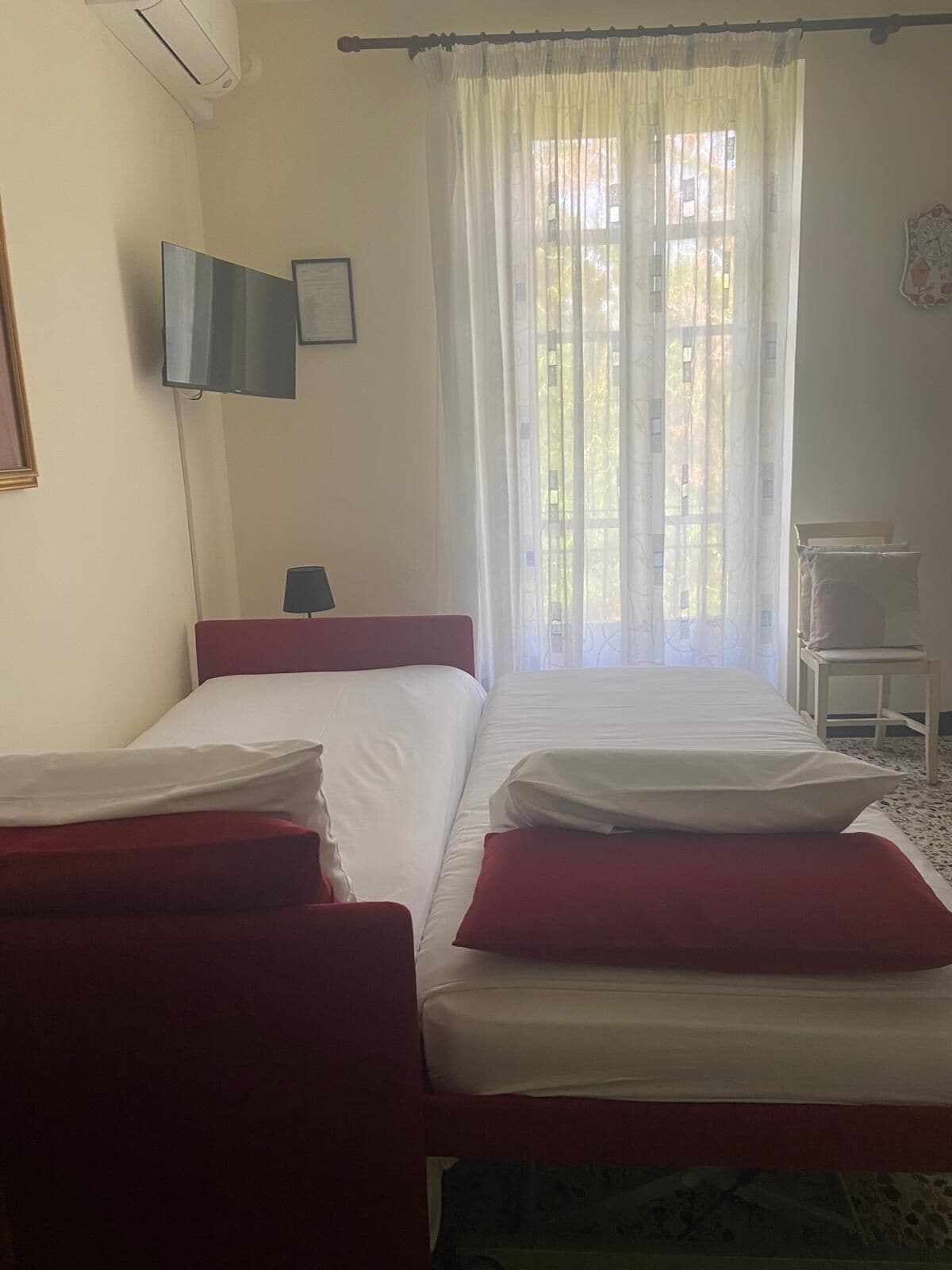 A hotel room with two beds, a TV, a window with sheer curtains, and a small table with a chair.