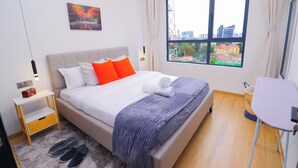 A modern hotel room with a large bed, a small bedside table, a window with a city view, and a desk with a chair.