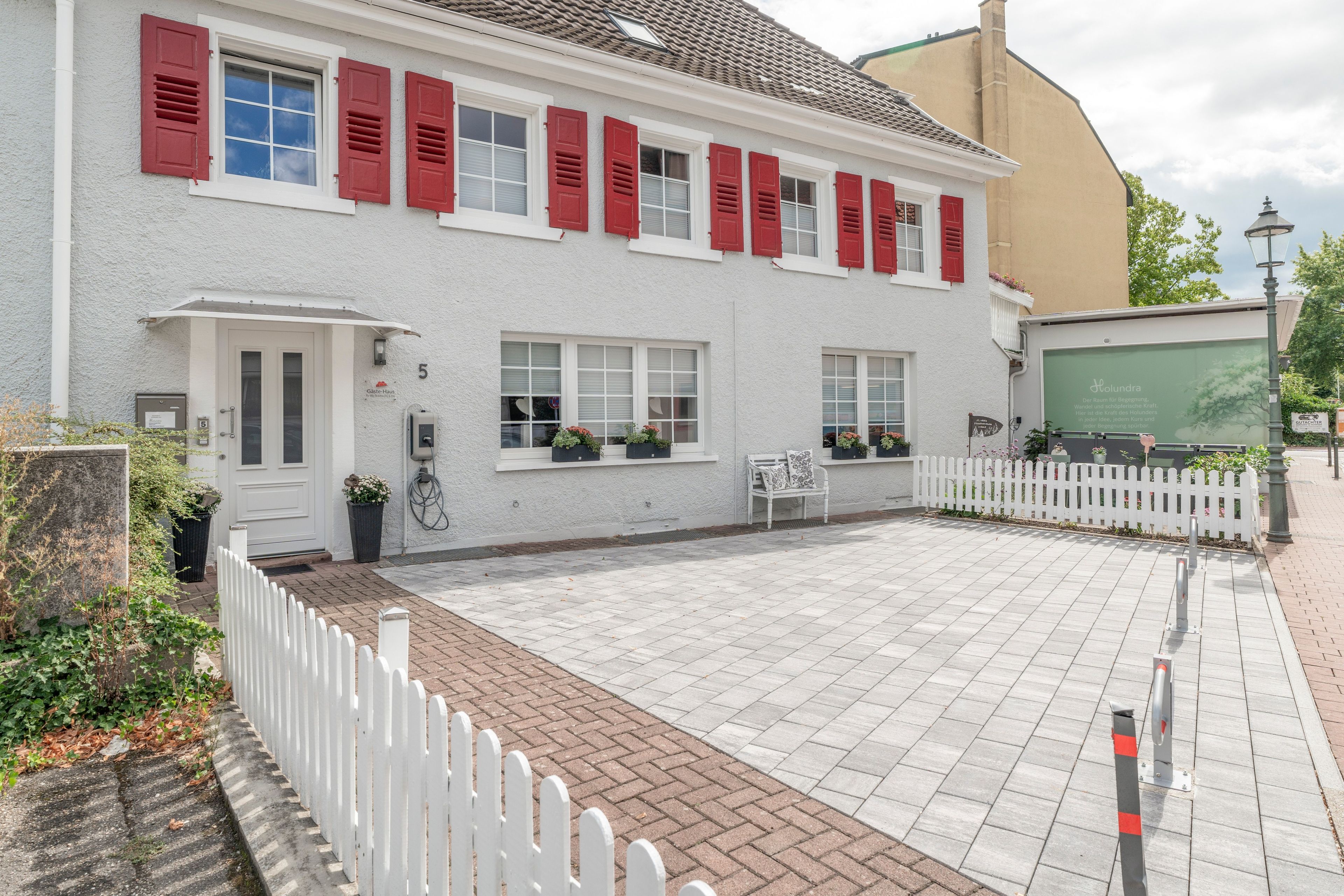 Ein weißes Gebäude mit roten Fensterläden, einem gepflasterten Innenhof und einem weißen Ziergartenzaun.