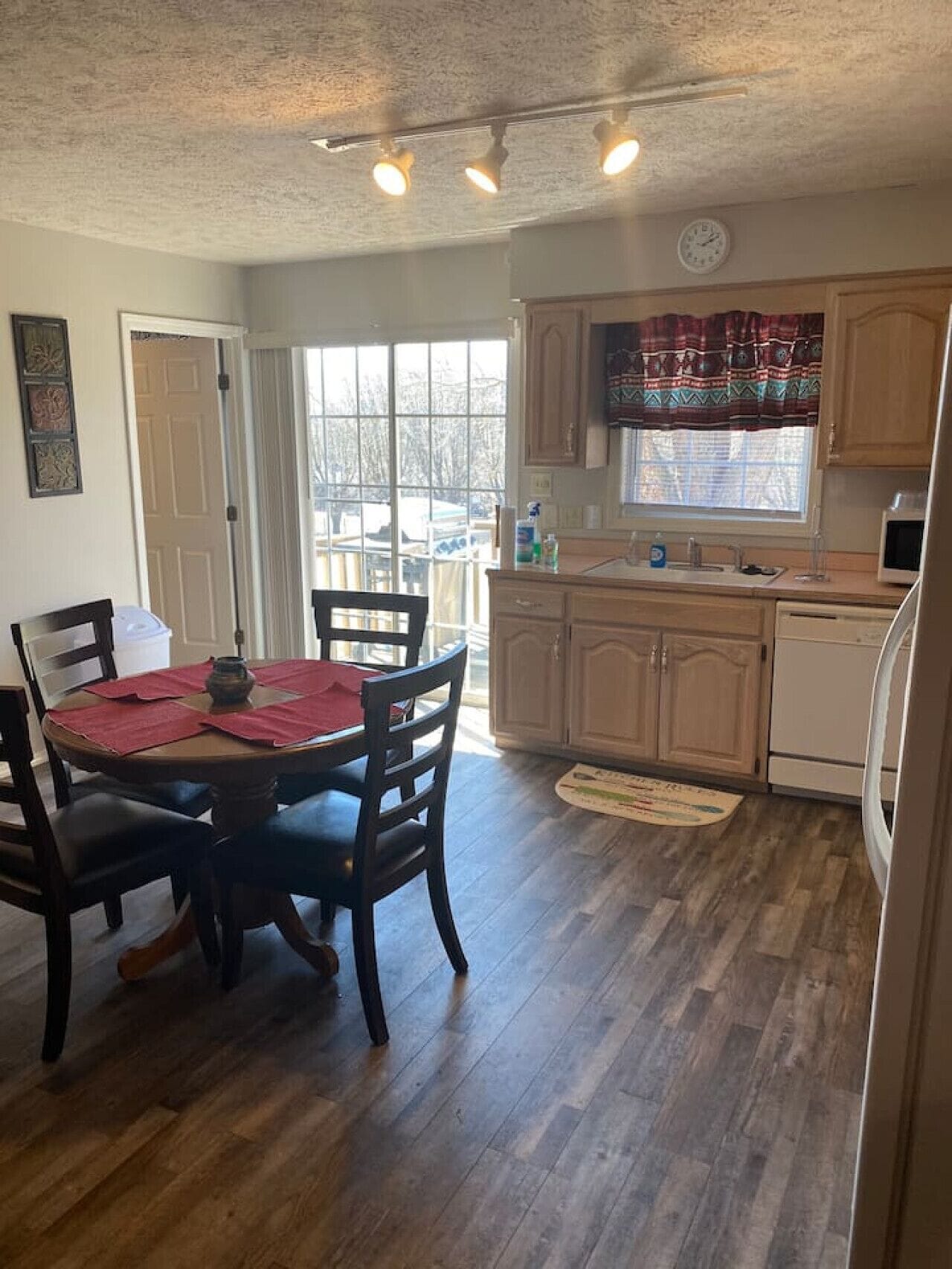 A dining area with a wooden table and chairs, a kitchen with wooden cabinets and a window, and a door leading outside.