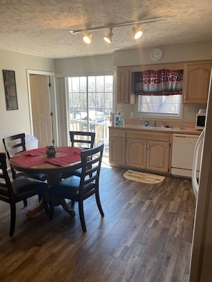 A dining area with a wooden table and chairs, a kitchen with wooden cabinets and a window, and a door leading outside.