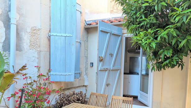 A wooden table and chairs outside a building with a blue shuttered door, a potted plant, and a small garden area.