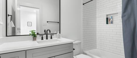 Une salle de bain moderne avec des carreaux blancs, un grand miroir, un lavabo blanc, des toilettes et une baignoire.