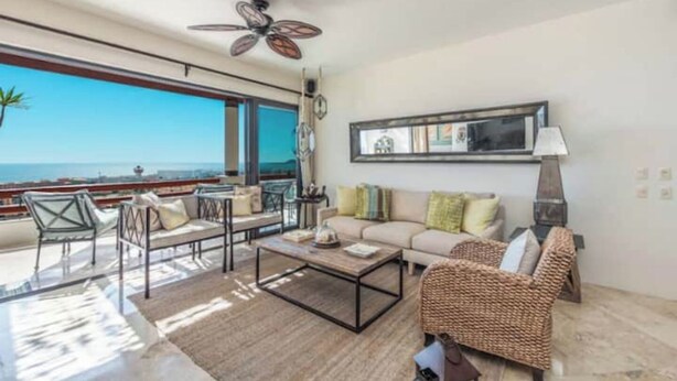 Ein geräumiges Wohnzimmer mit großem Fenster und Meerblick, Deckenventilator, einer Couch mit Kissen und einem Couchtisch.