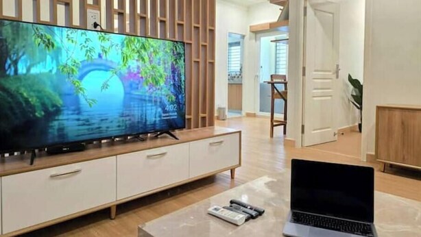 A modern living room with a large flat-screen TV on a wooden cabinet, a laptop on a marble coffee table, and a door leading to another room.