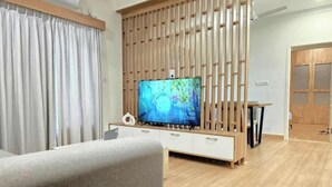 A modern living room with a flat-screen TV, wooden panel wall, and a dining area with a table and chairs.