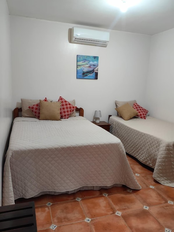 A room with two beds, each with a quilted bedspread and patterned pillows, a painting on the wall, and an air conditioning unit above the beds.