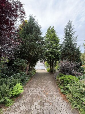 Un chemin pavé mène à un bâtiment blanc doté d’une fenêtre, qui est entouré d’un feuillage dense et d’arbres.