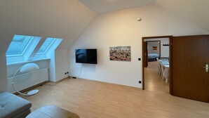 A spacious room with a sloped ceiling, a flat-screen TV mounted on the wall, a painting, and a door leading to another room. - Cozy vacation apartment on the edge of the forest - ideal for nature lovers & families (Bark)