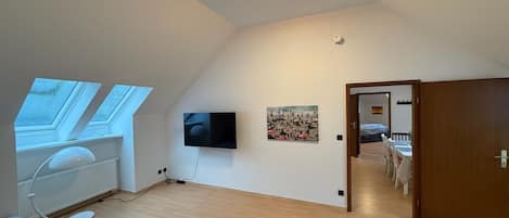 Een ruime kamer met een schuine plafond, een flatscreen televisie aan de muur, een schilderij en een deur naar een andere kamer.