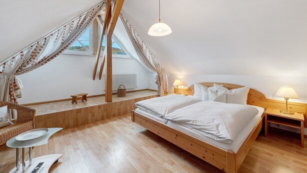 Un dormitorio con una cama de madera, una mesita, una silla, una lámpara y un ventanal con cortinas.