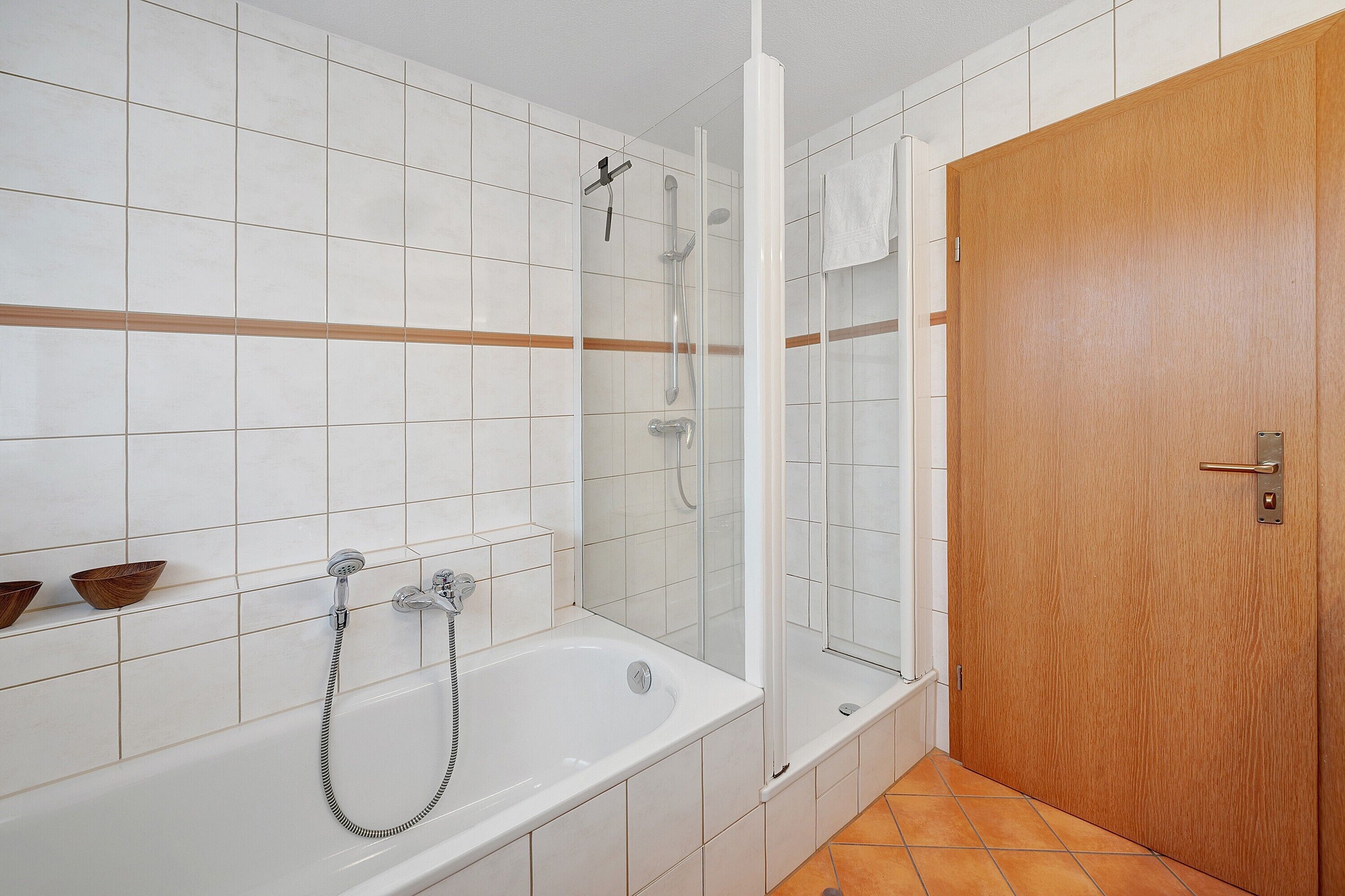 Une salle de bain avec des murs recouverts de carreaux blancs, une porte en bois, une baignoire et un espace douche.