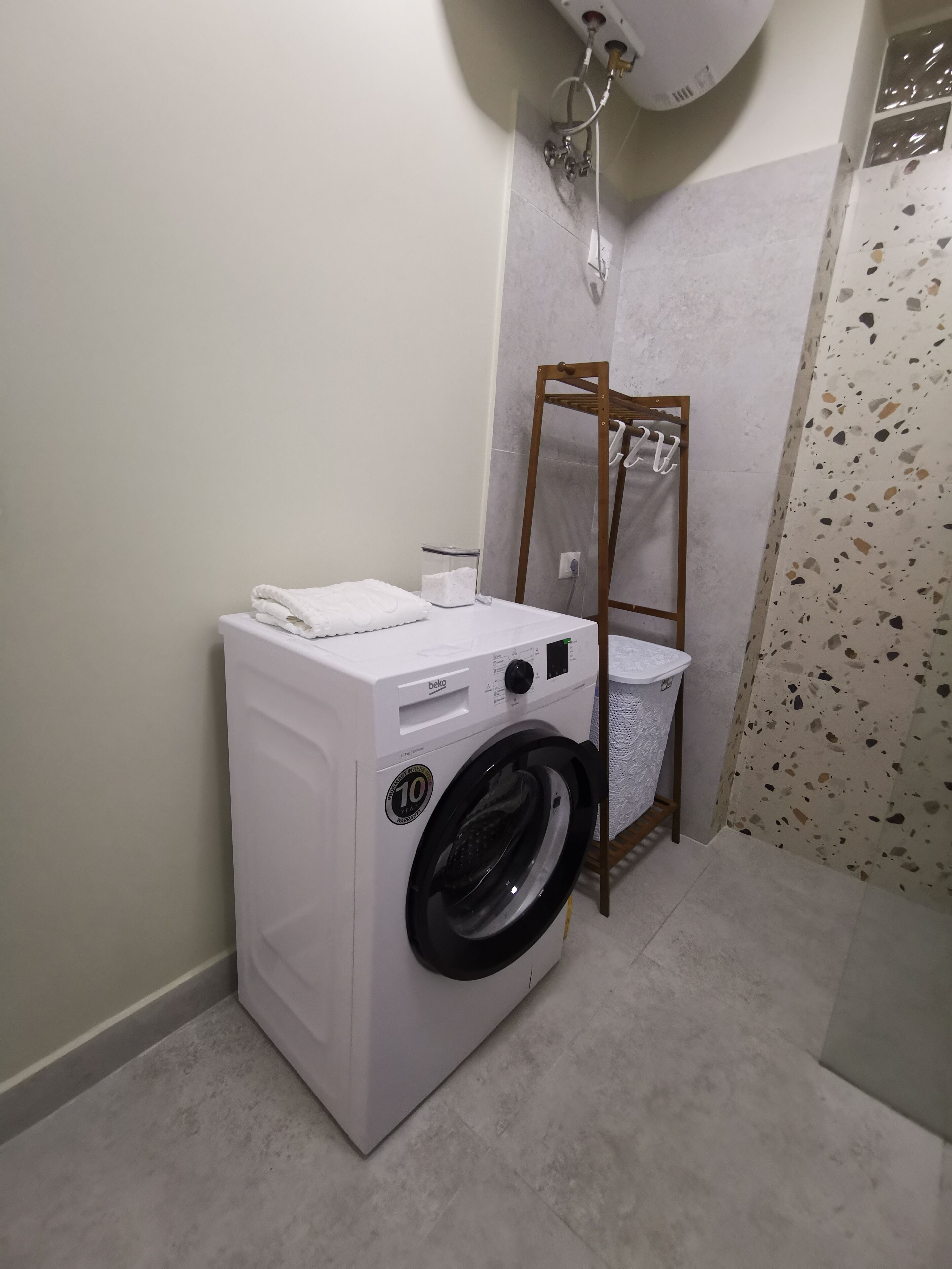 Un espace réduit dédié à la buanderie, comprenant un lave-linge à chargement frontal, un étendoir et une petite poubelle.