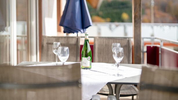 A table with a bottle of wine and two glasses, a blue umbrella, and a view of a body of water and buildings in the background. - Holiday Apartment 'Landblick' with Balcony and Wi-Fi (Biederbach)