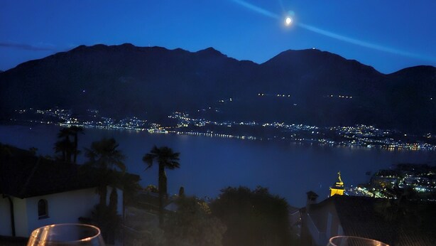 Una mesa con copas de vino y velas, con vistas a un lago con luces de la ciudad y montañas al fondo.