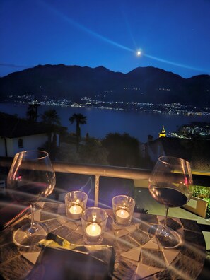 A table with wine glasses and candles, overlooking a lake with city lights and mountains in the background.
