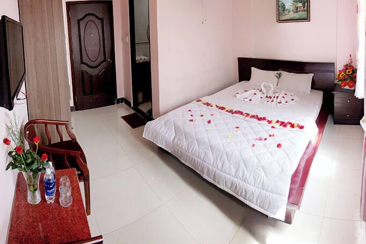 A neatly arranged bedroom with a bed adorned with rose petals, a wooden chair, and a small table with decorative items.