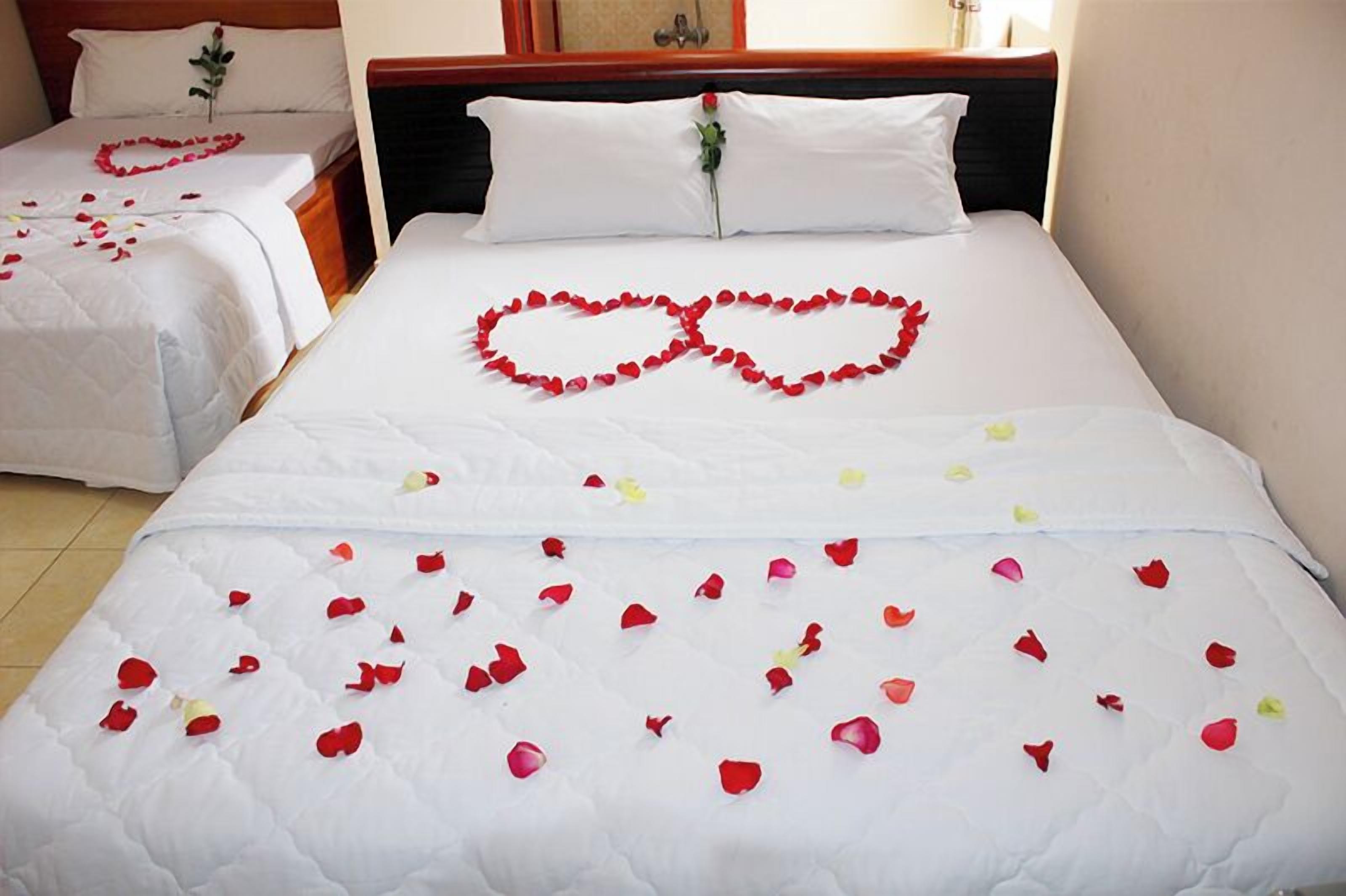 A hotel room with two beds, one with a heart-shaped arrangement of rose petals and another with a simple rose on the headboard.
