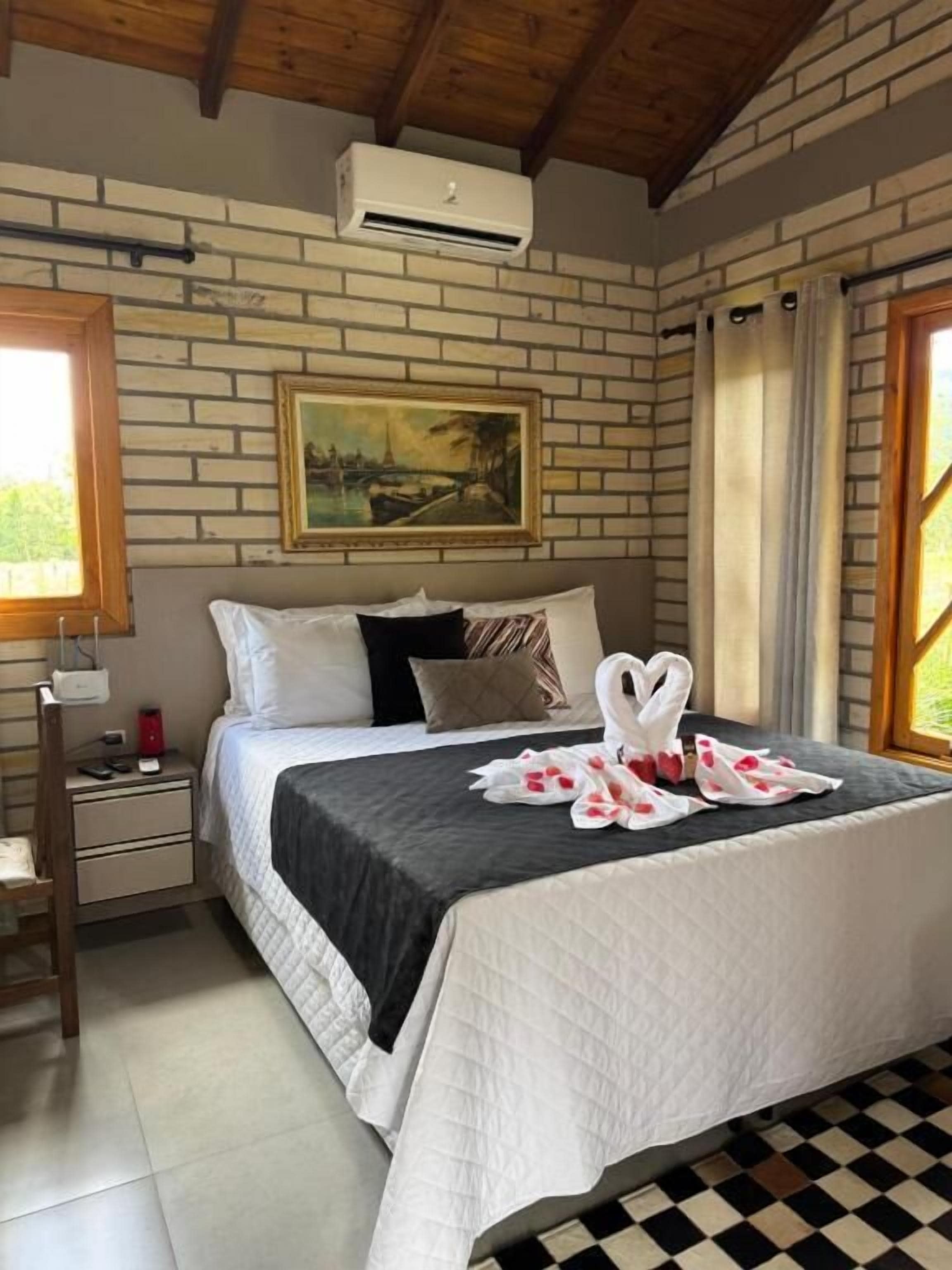 A hotel room with a brick wall, a painting, a bed with a black bedspread, a nightstand with a lamp, and a checkered floor.