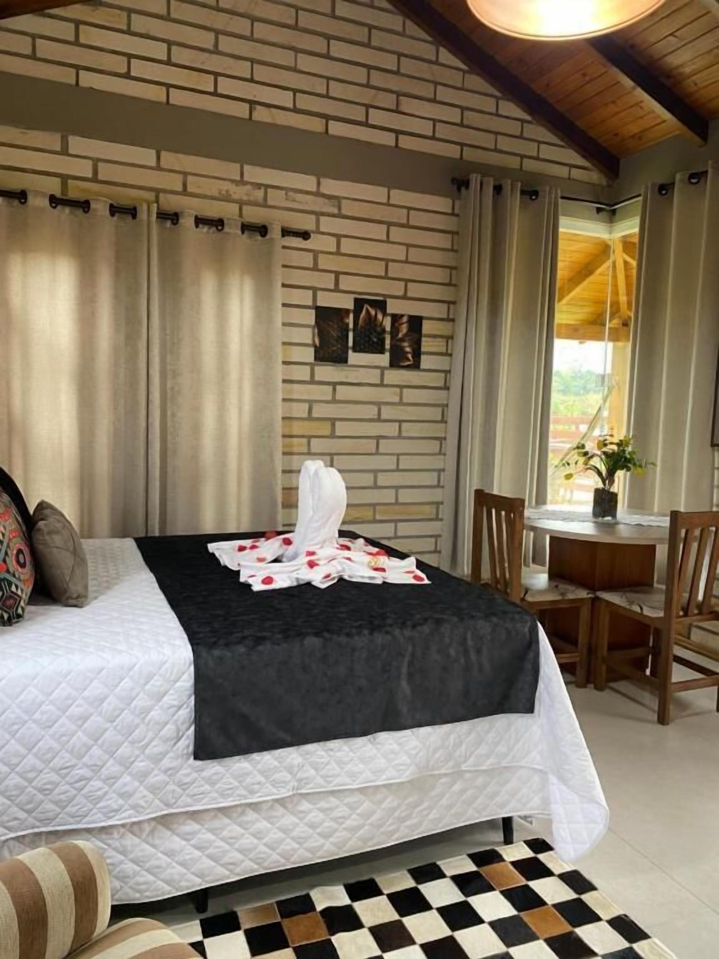 A hotel room with a brick wall, a bed with a black and white quilt, a dining area with a table and chairs, and a window with curtains.