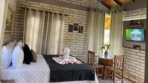 A hotel room with a brick wall, a bed with white and black bedding, a TV on the wall, and a small table with chairs. - Pousada Mirante dos Canyons (Praia Grande)