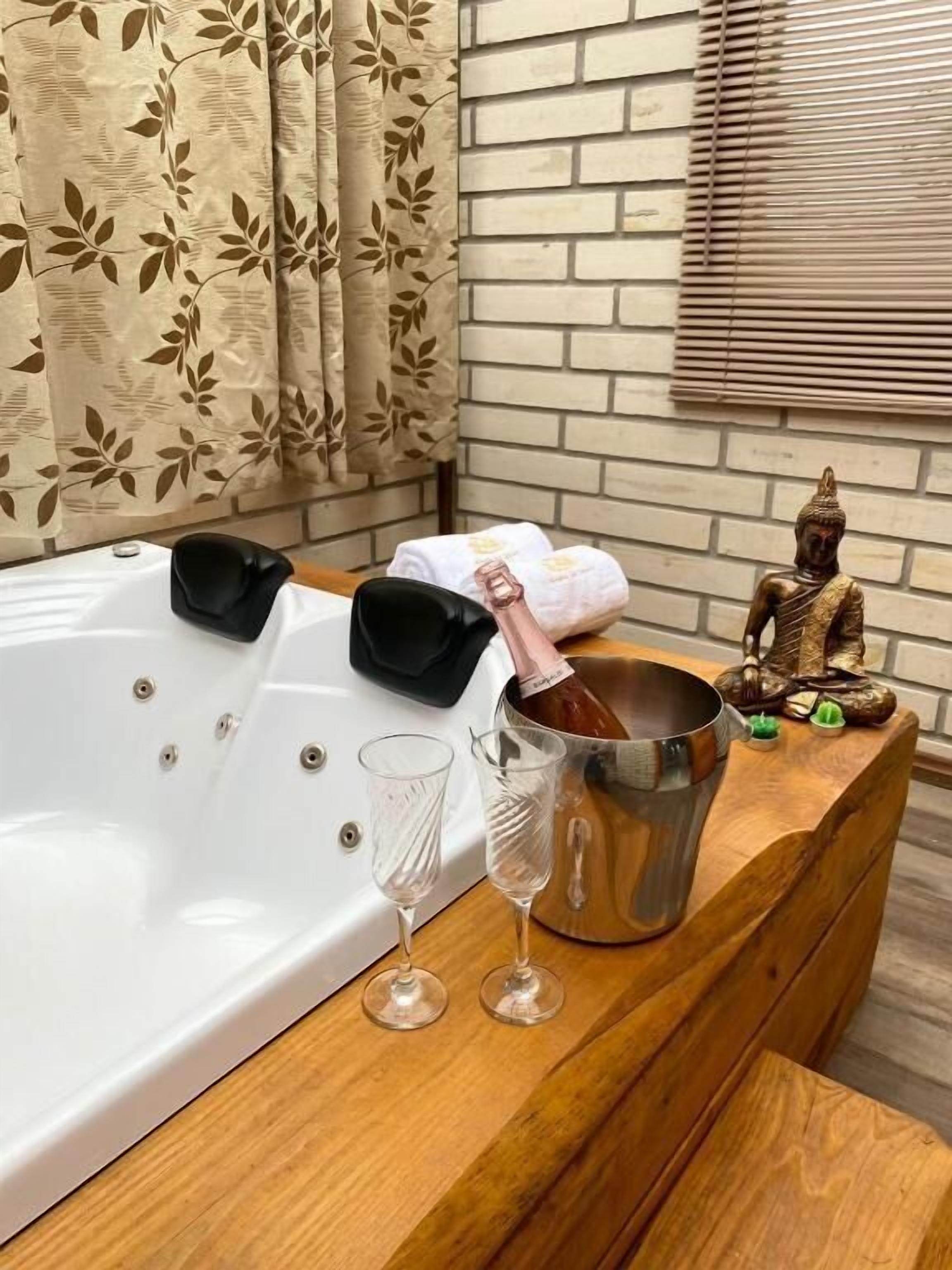 A bathroom with a white bathtub, two black bathrobes, a bucket with a bottle, and two wine glasses on a wooden vanity.