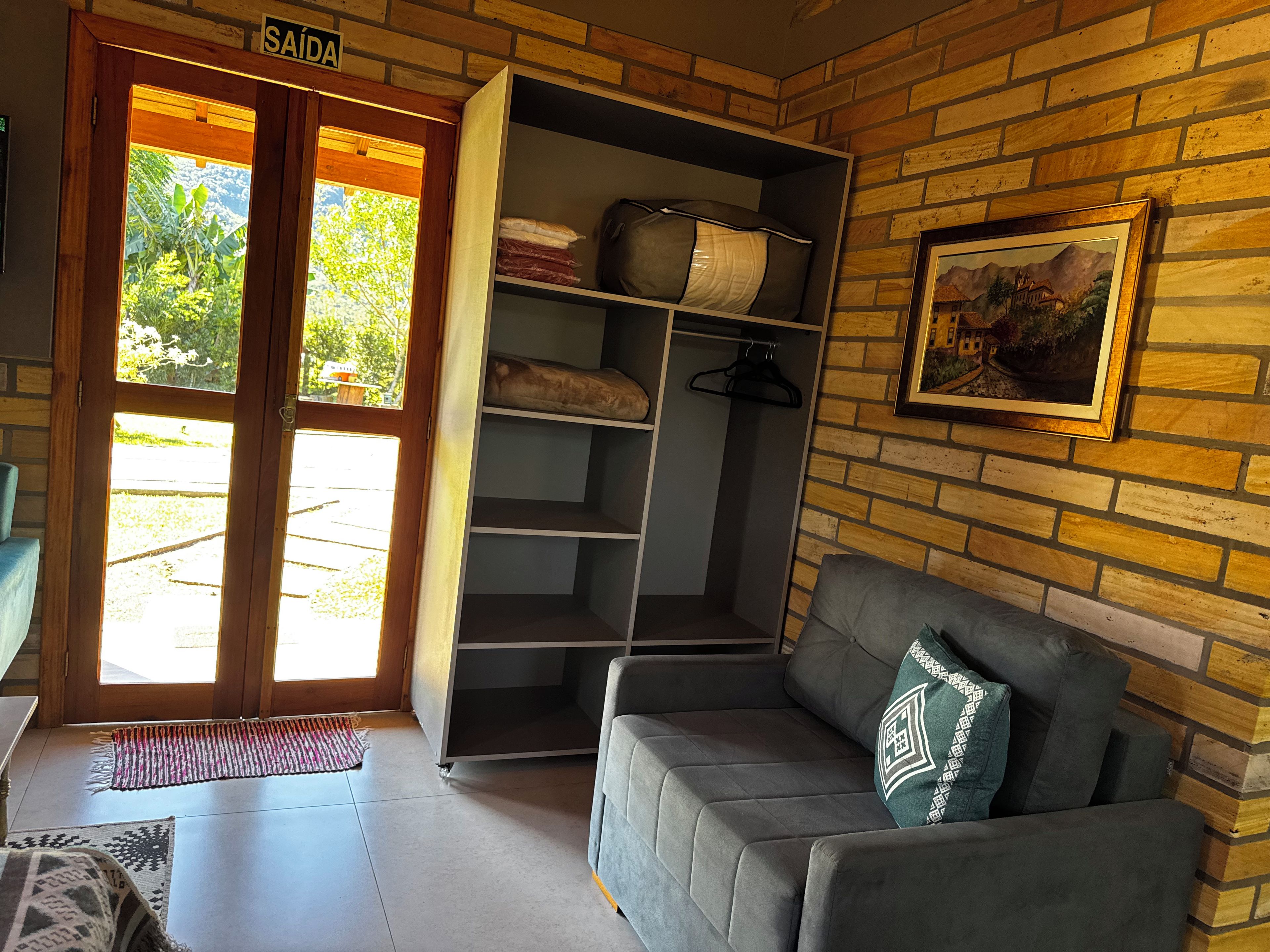 Una habitación con un sofá gris, un armario con estantes, una puerta que da al exterior y un cuadro en la pared.