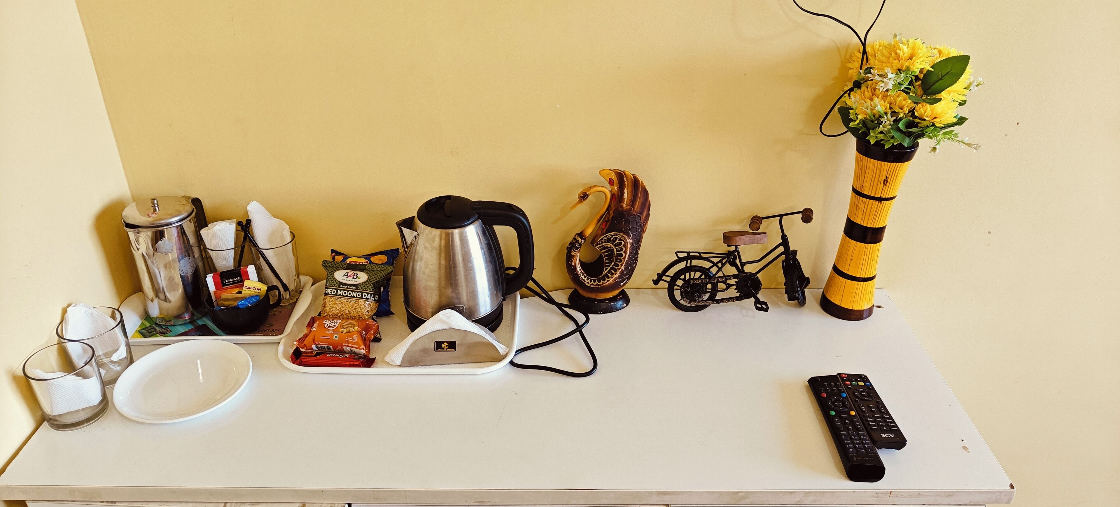 A kitchen counter with a coffee maker, a kettle, a small bicycle, a vase with flowers, a remote control, and some food items.