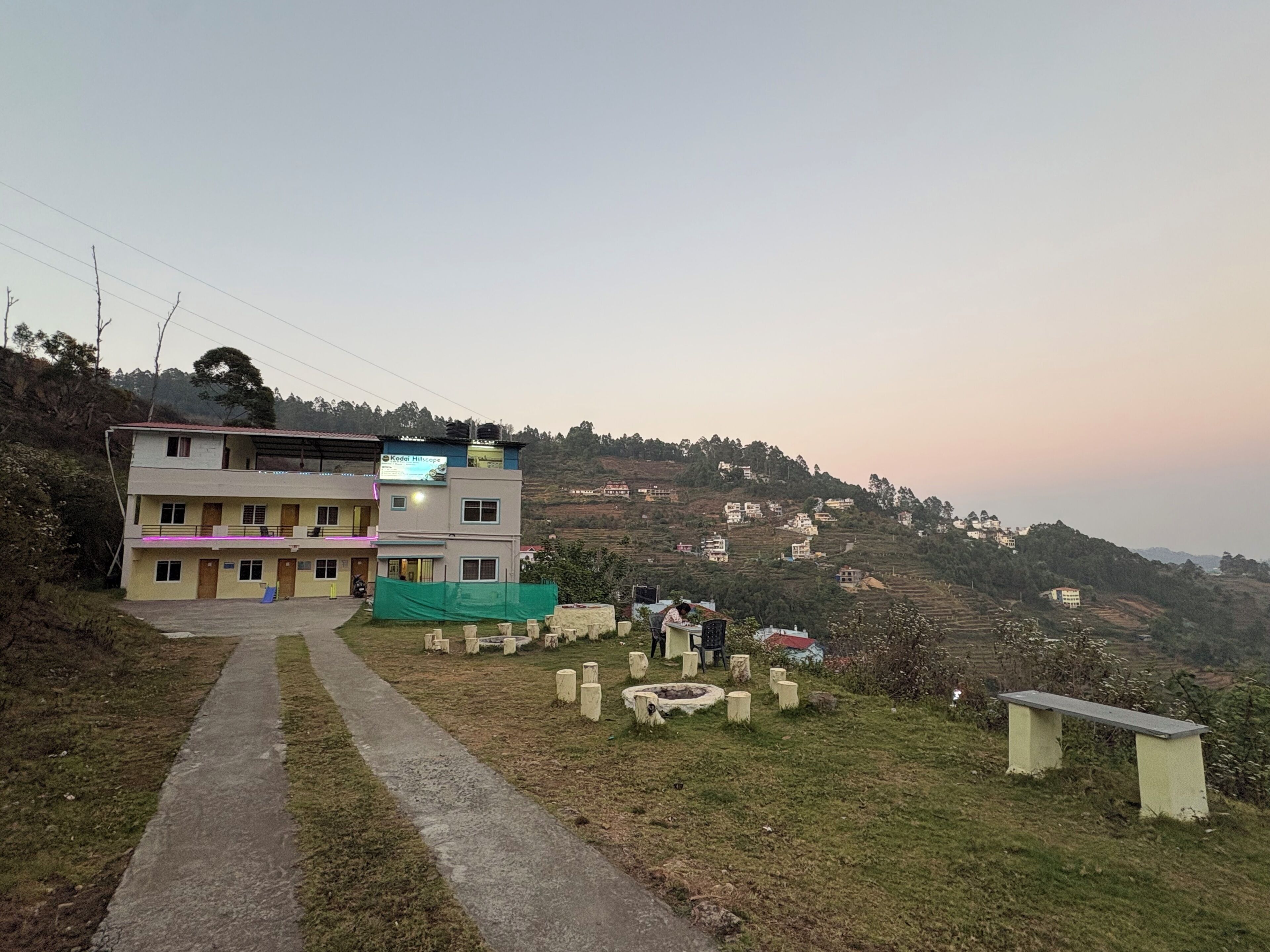 A multi-story building with a flat roof, a paved pathway leading to the entrance, and a hilly landscape in the background.