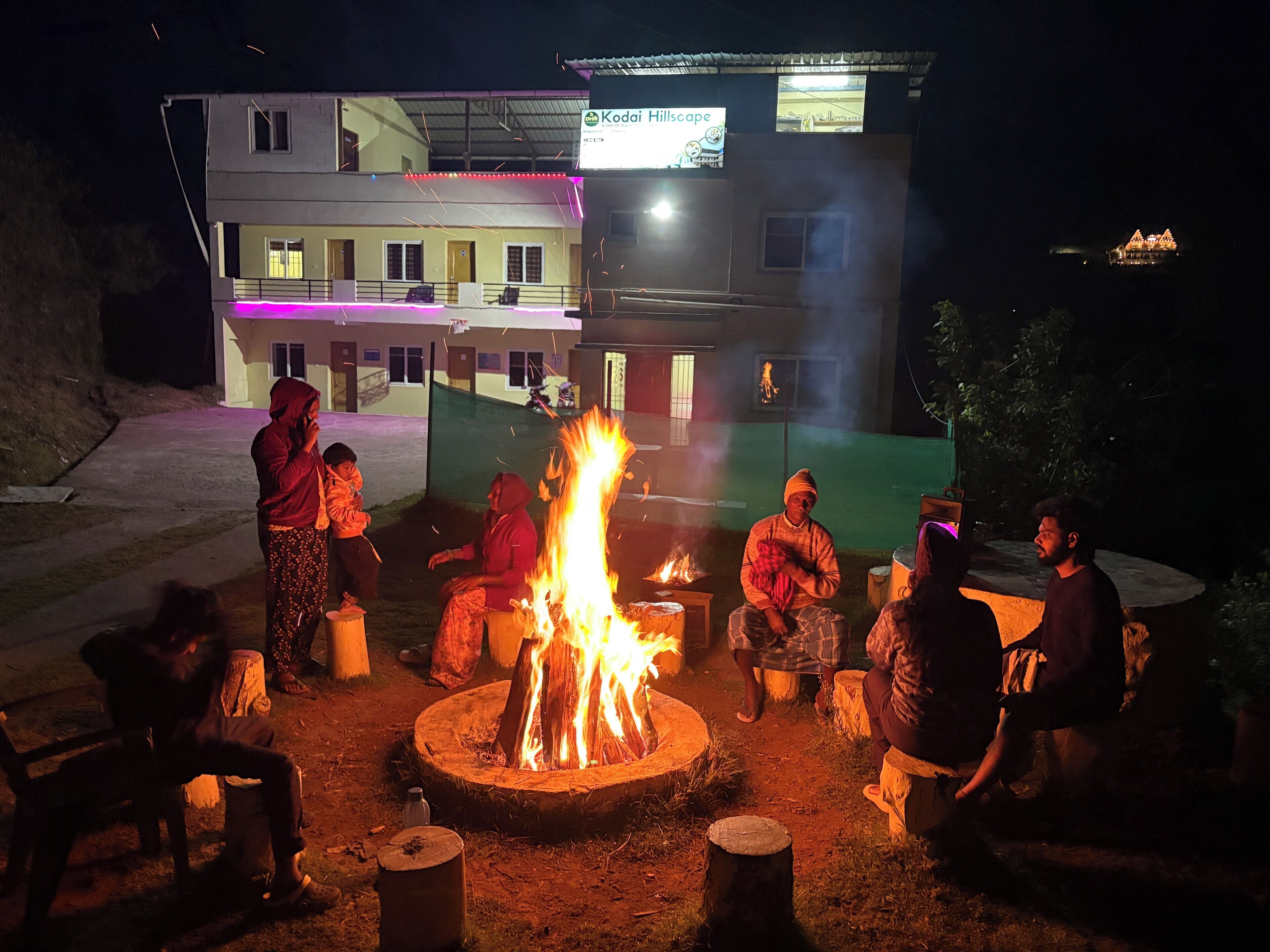 A group of people around a campfire at night, with a building in the background.
