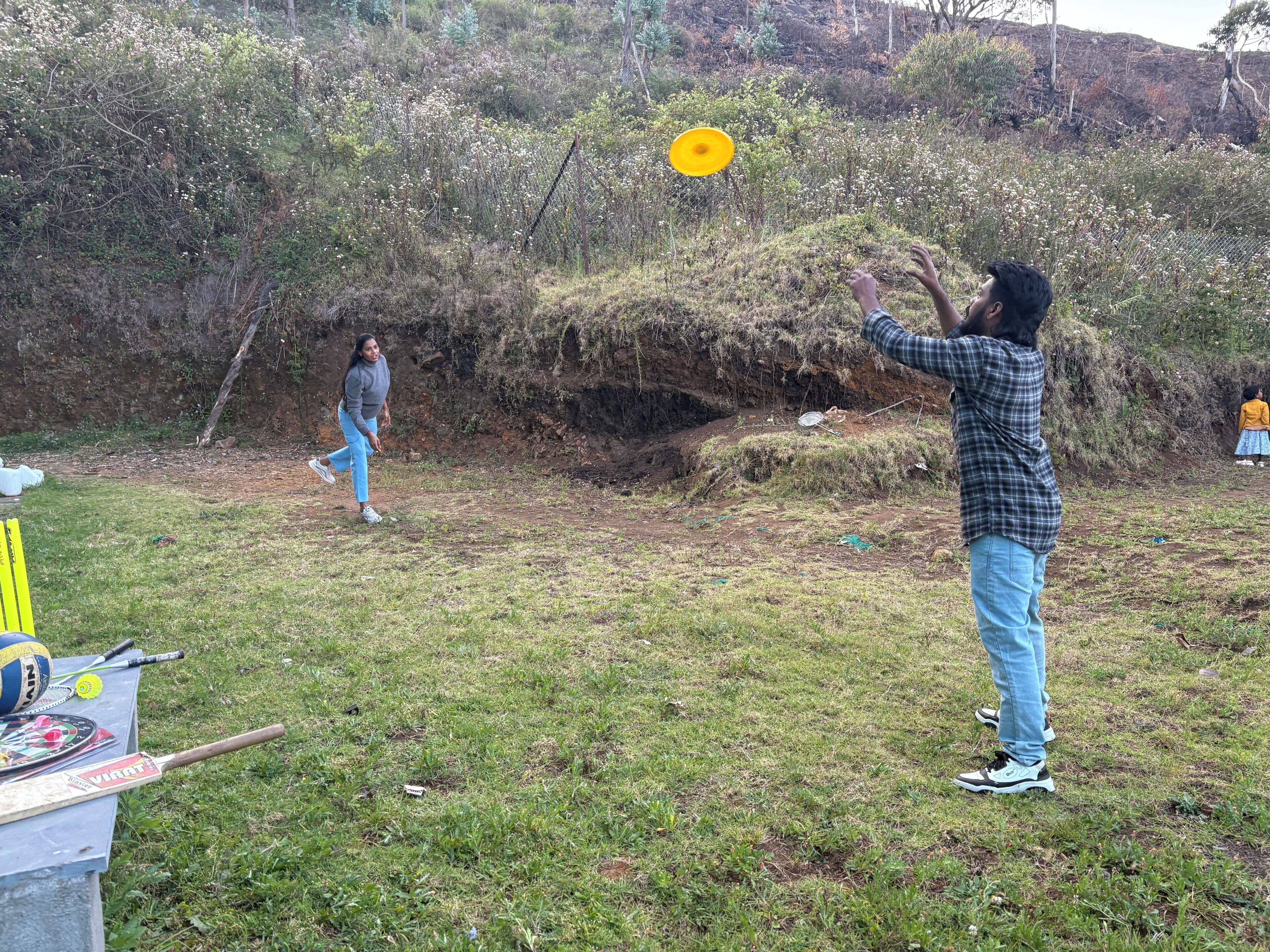 An outdoor area with grass, a table with sports equipment, and a person playing with a yellow frisbee.