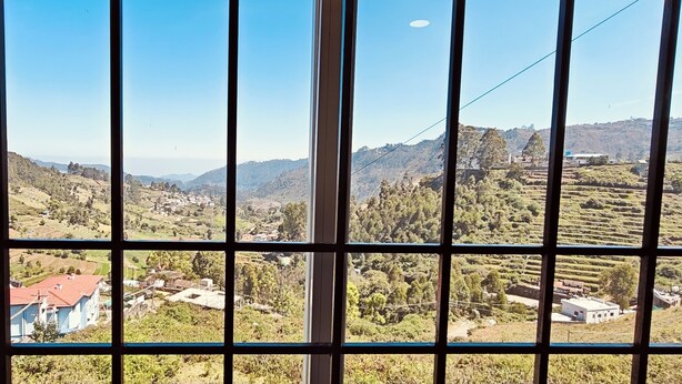 A view through a window with a grid pattern, displaying a rural landscape with terraced fields, scattered houses, and hills in the distance. - Kodai Hillscape (Kodaikanal)