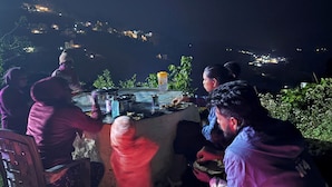 A group of people gathered around a table with cooking equipment, seated on chairs, outdoors at night with a view of a hillside town in the background. - Kodai Hillscape (Kodaikanal)
