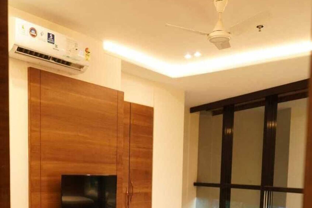 A modern bedroom with a bed, a TV mounted on a wooden cabinet, an air conditioning unit, and a window with blinds.