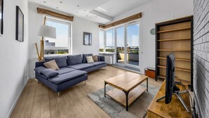 Un salon moderne avec un canapé, une table basse et un téléviseur. Il y a de grandes fenêtres et un balcon.