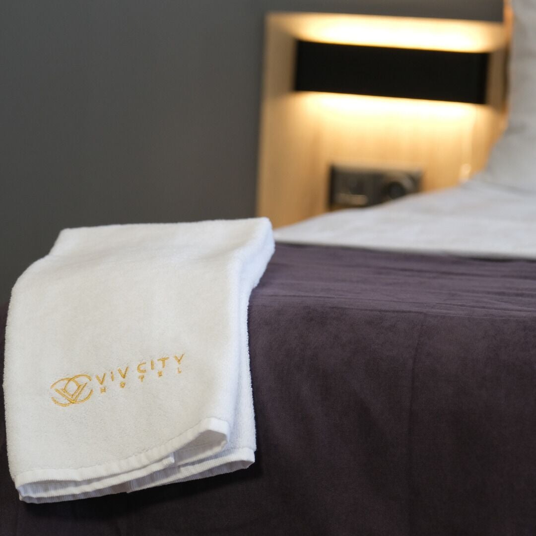 A neatly folded white towel with "VIVICITY HOTEL" embroidered on it, placed on a bed with a purple bedspread.