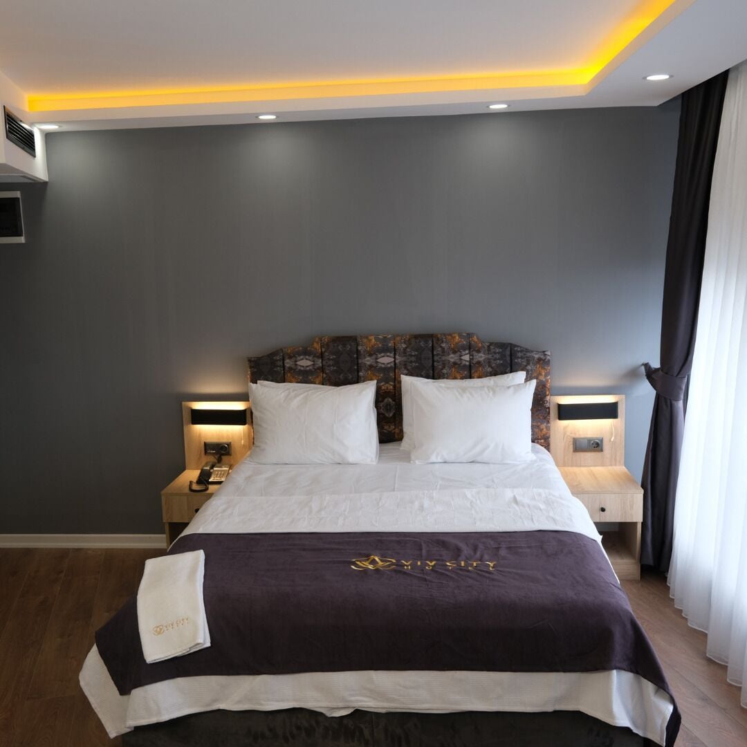 A hotel room with a large bed, two bedside tables with lamps, a headboard with a patterned design, and a dark-colored wall.