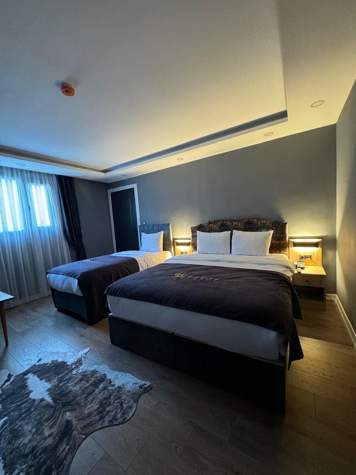 A hotel room with two beds, a wooden floor, a large rug, and a nightstand with a lamp.