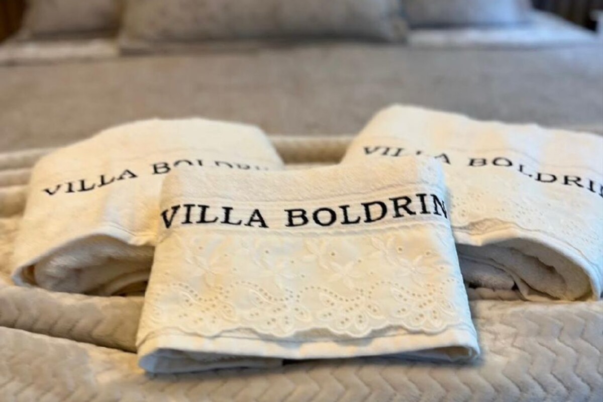 Un lit recouvert d’une couvre-lit beige à motifs et deux serviettes pliées sur lesquelles est imprimé le nom « Villa Boldrin ».