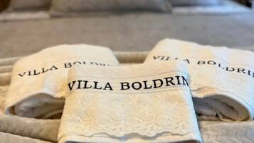Cama com colcha bege estampada e duas toalhas dobradas com a inscrição "Villa Boldrin".