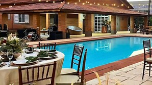 Ein Essbereich am Pool mit einem Tisch für zwei Personen, einer überdachten Lounge-Ecke und einem Gebäude mit roten Ziegeldächern.