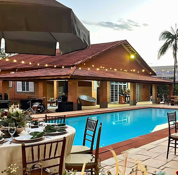 Un espace repas aménagé au bord de la piscine, avec une table dressée pour deux personnes, un coin salon abrité et un bâtiment surmonté d’un toit recouvert de tuiles rouges.