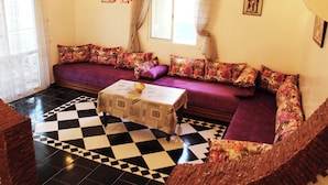 Ein Wohnzimmer mit Schachbrettmuster-Boden, einer Couch mit gemusterten Kissen, einem Tisch mit einer verzierten Tischdecke und einer Kandelaber.